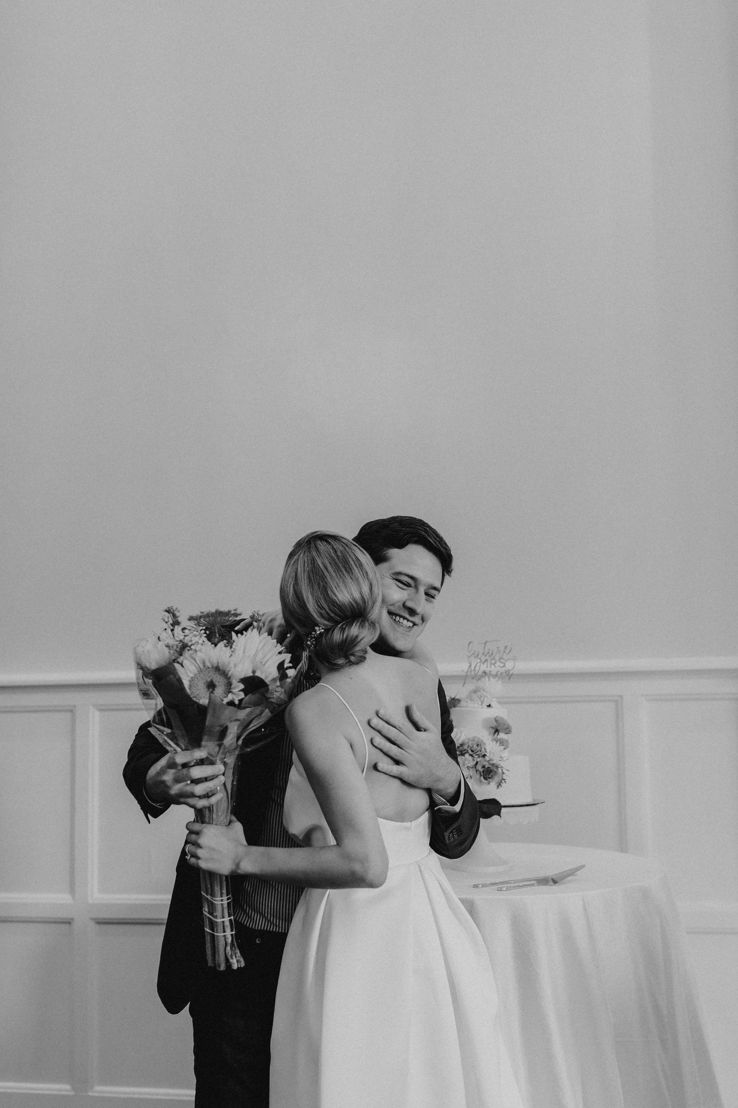 Black and white candid moment of newlyweds embracing while holding flowers and champagne.