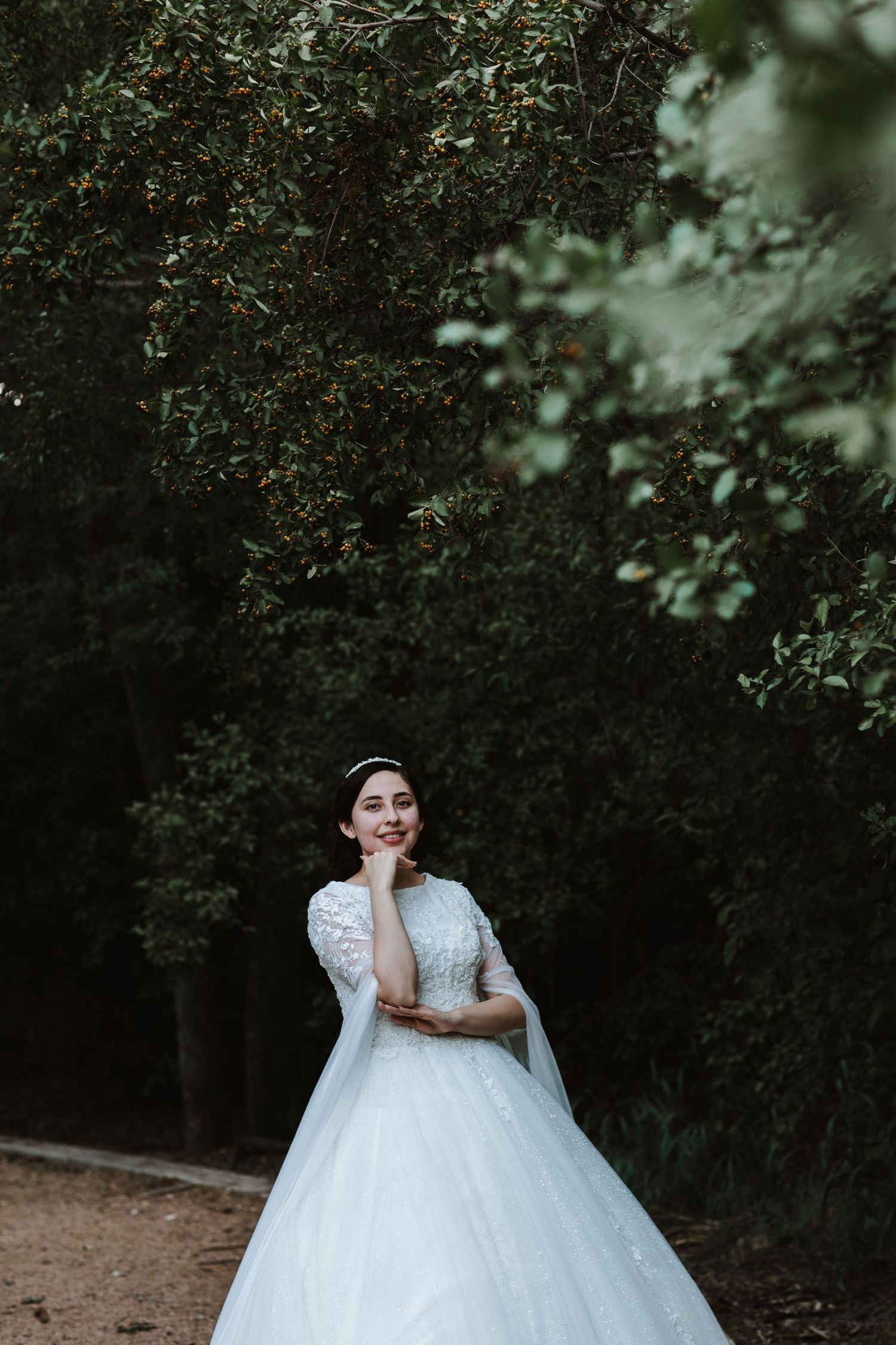 Bride in a white ball gown with lace sleeves posing among lush green trees.
