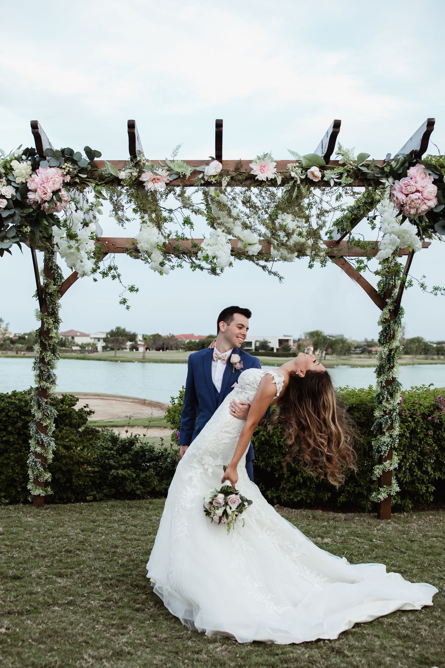 Bride and groom share romantic moment under floral wooden arch with pink flowers and greenery beside a lake.