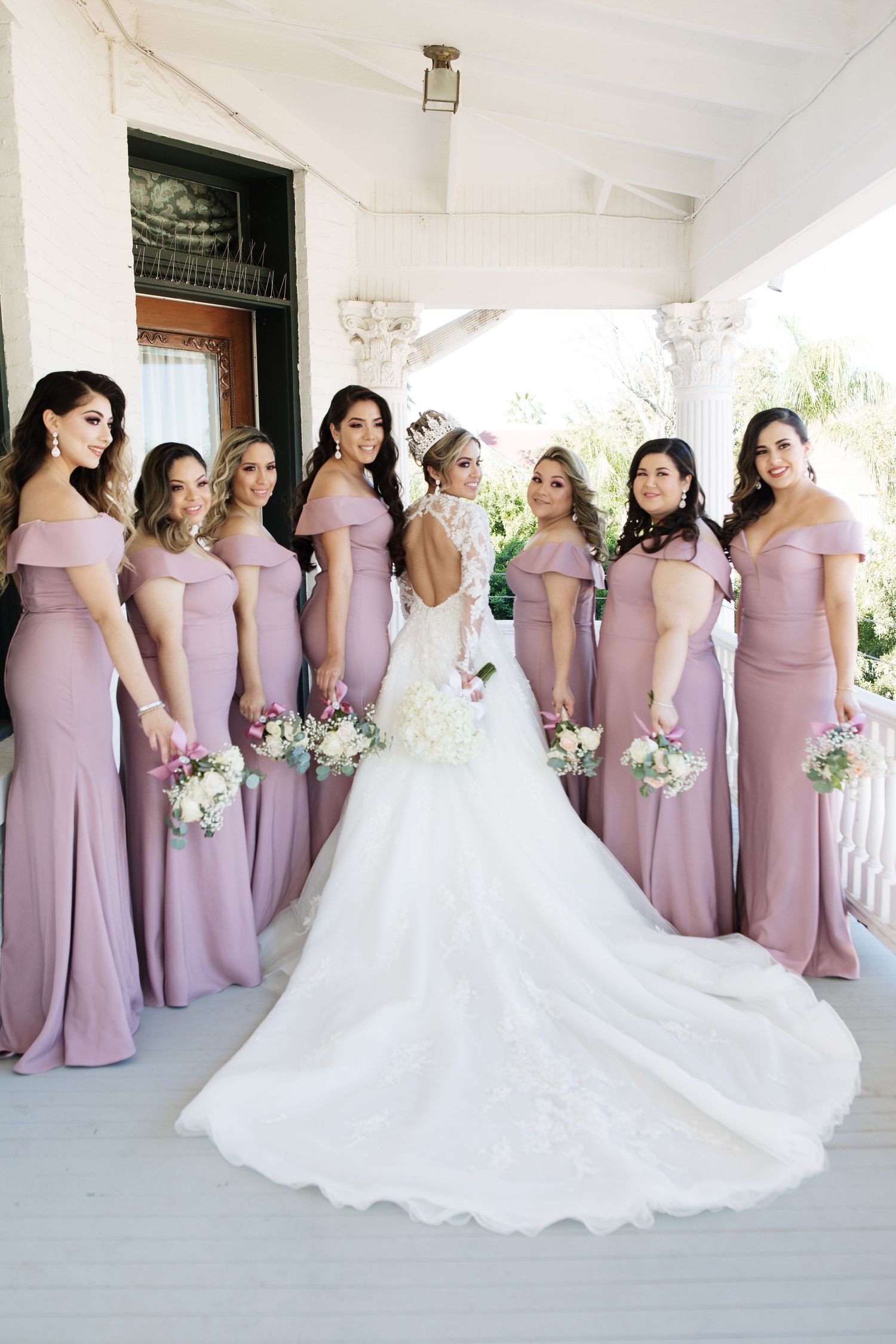 Bride in lace gown poses with six bridesmaids in mauve off-shoulder dresses holding bouquets on porch.