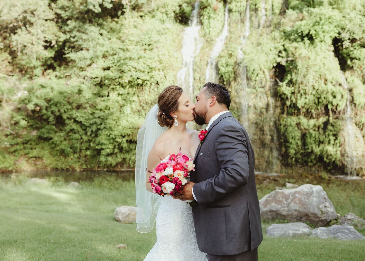 Bride and groom sharing a kiss near a waterfall backdrop holding a red bridal bouquet.