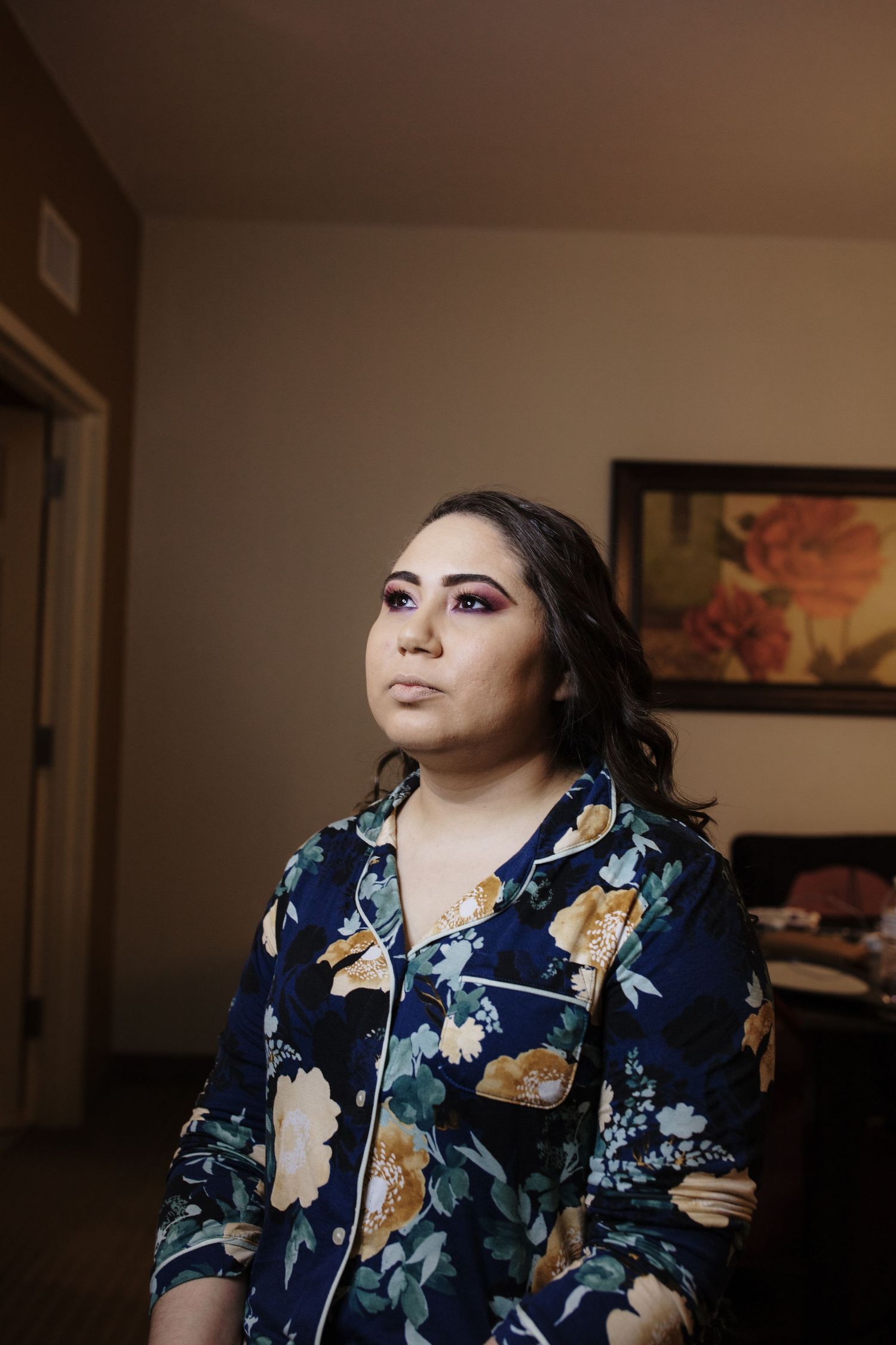 Woman in floral blue pajamas with bold purple eye makeup gazes upward in a softly lit room.