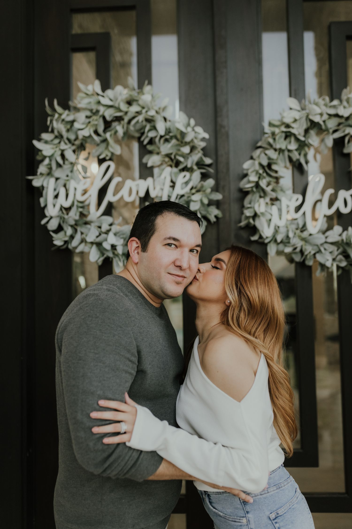 A couple poses romantically in front of decorative welcome wreaths, woman kissing mans cheek while embracing outdoors.