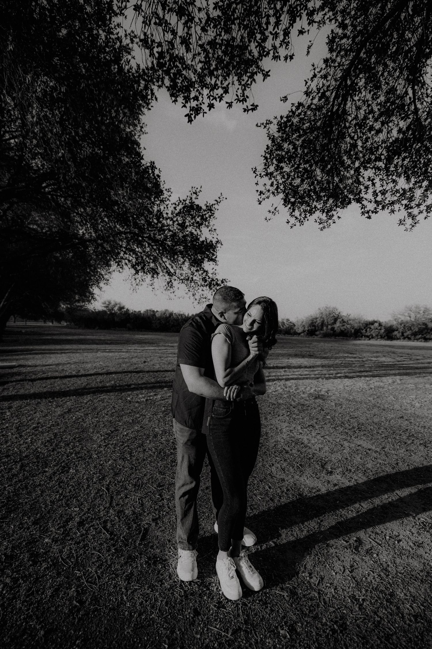 A couple embraces tenderly under a large tree in a black and white outdoor engagement photo.