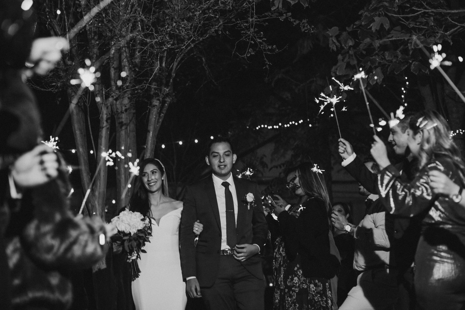 Bride and groom walking through a sparkler send-off at night surrounded by cheering wedding guests.