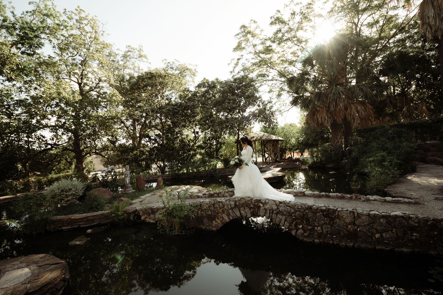 Bride in flowing white gown standing on a stone bridge surrounded by lush greenery and a pond.