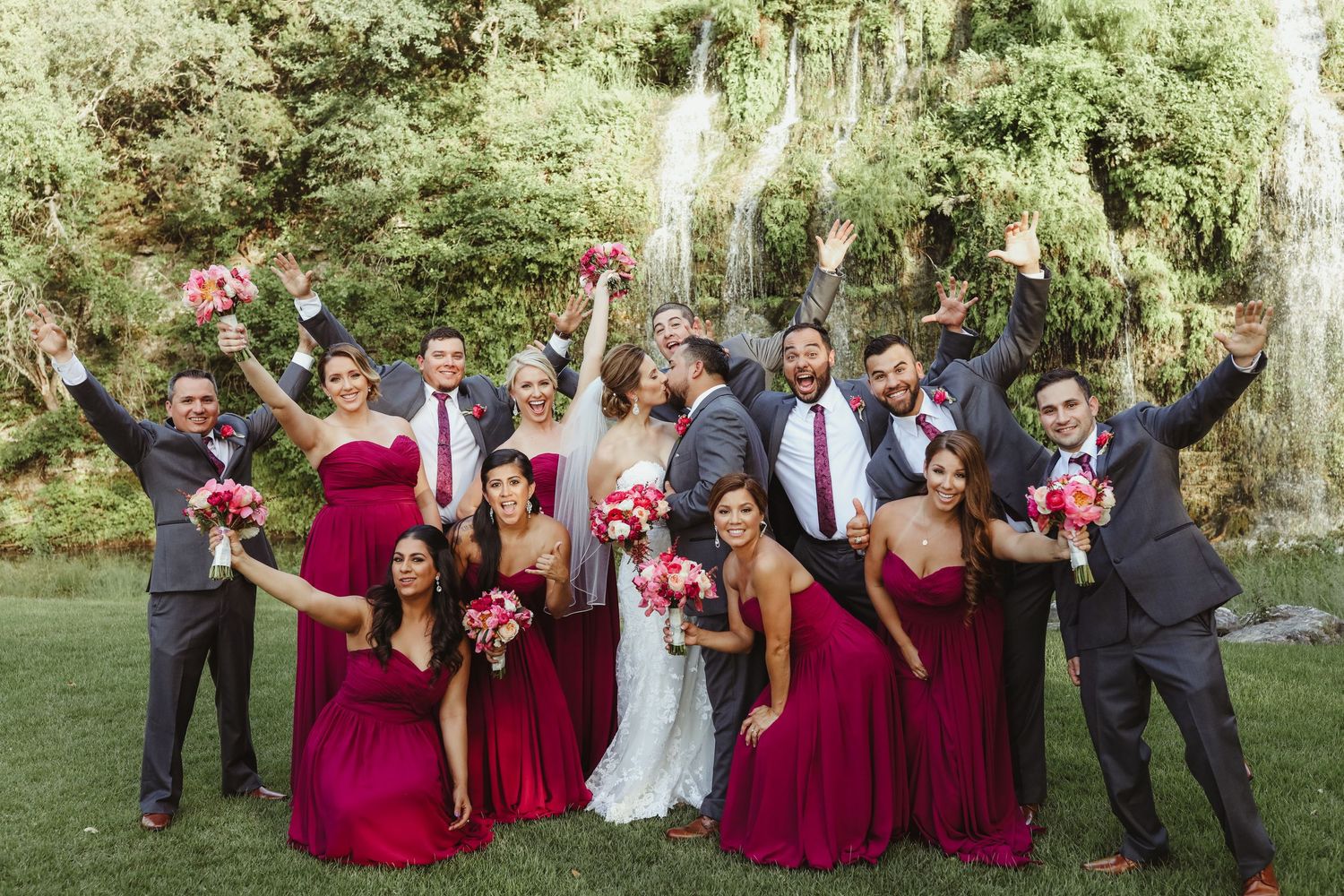 Exuberant wedding party in burgundy and gray celebrating in front of a waterfall outdoors.