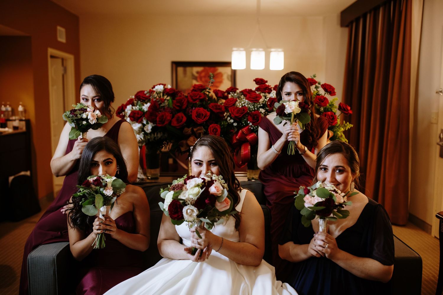 Bridesmaids in burgundy dresses hiding faces behind bouquets with bride in white at wedding preparation.