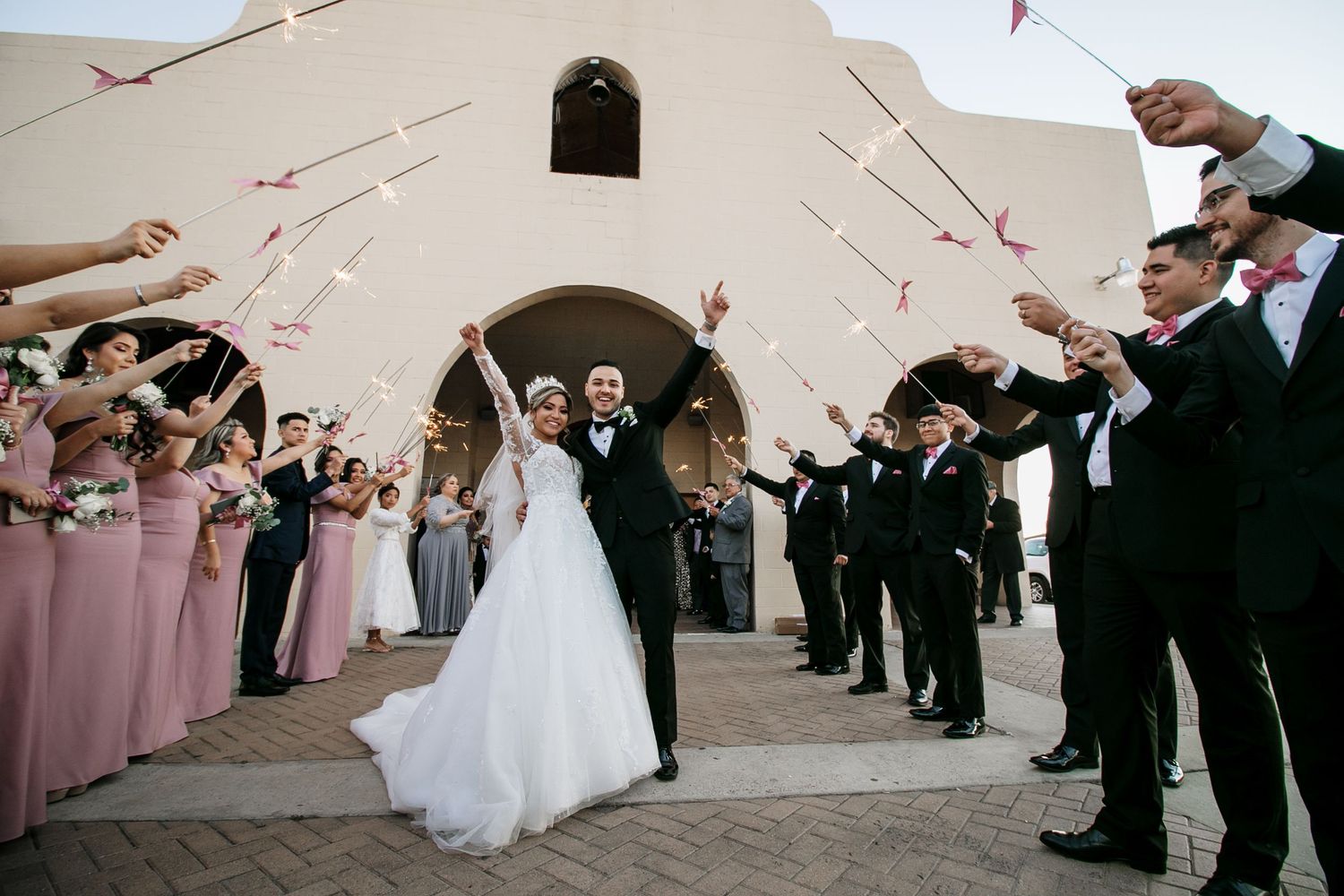 Newlyweds celebrate with wedding party as guests wave streamers outside a Spanish-style church.