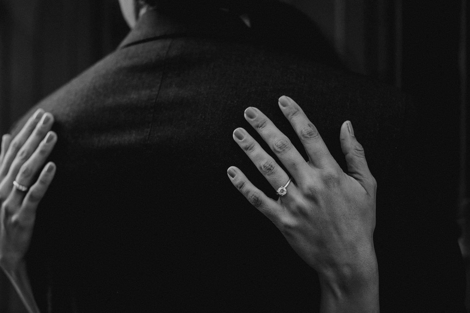 Close-up black and white photo of a woman's hands with an engagement ring resting on a man's dark jacket.