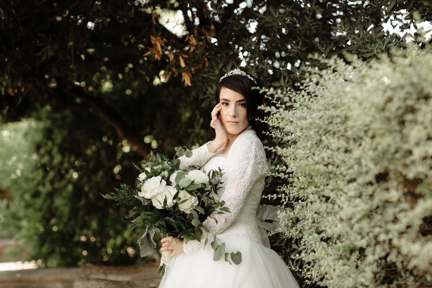 Elegant bride in lace wedding gown holds white floral bouquet surrounded by lush green trees.