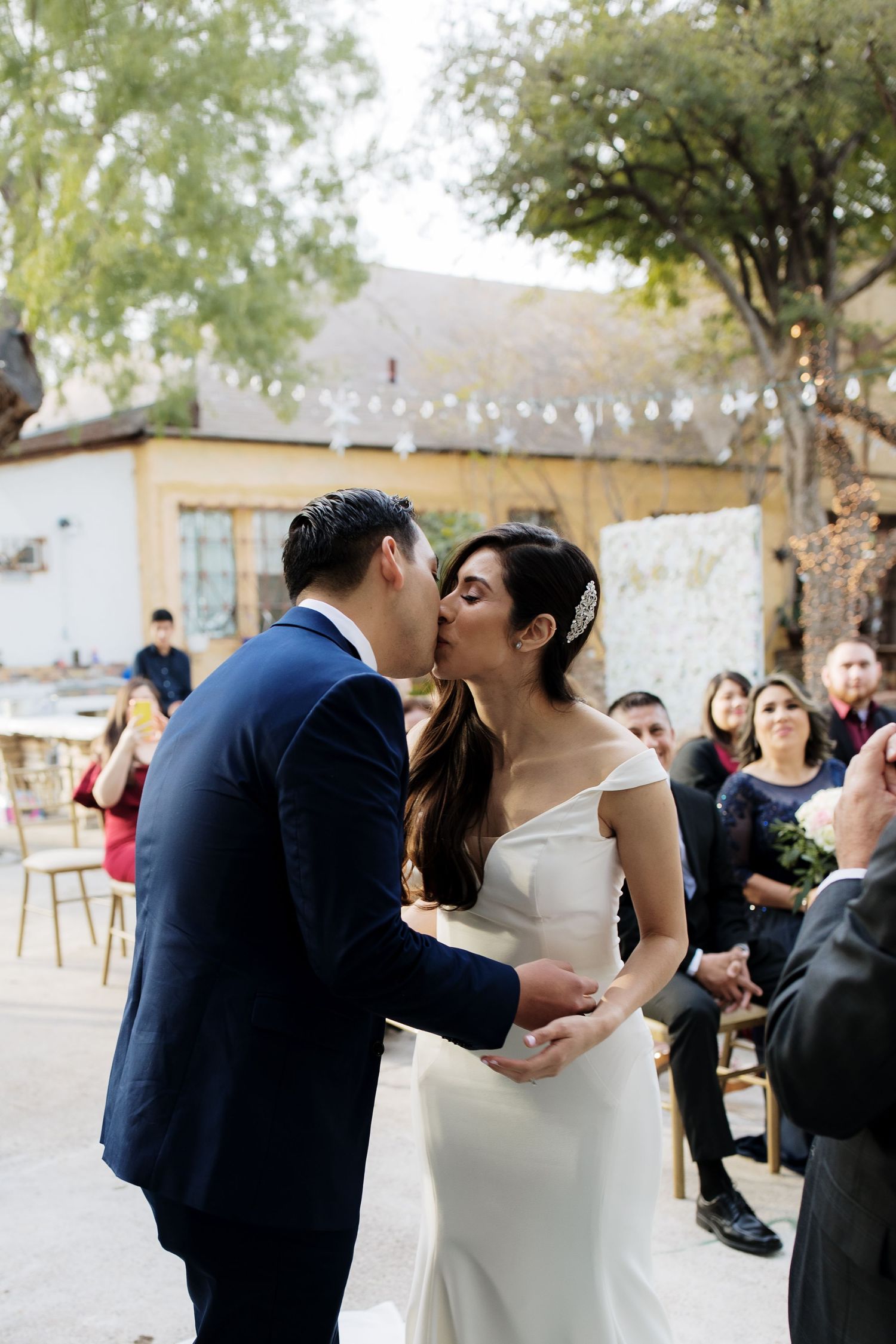 Bride and groom share their first kiss at an outdoor wedding ceremony with string lights in the background.