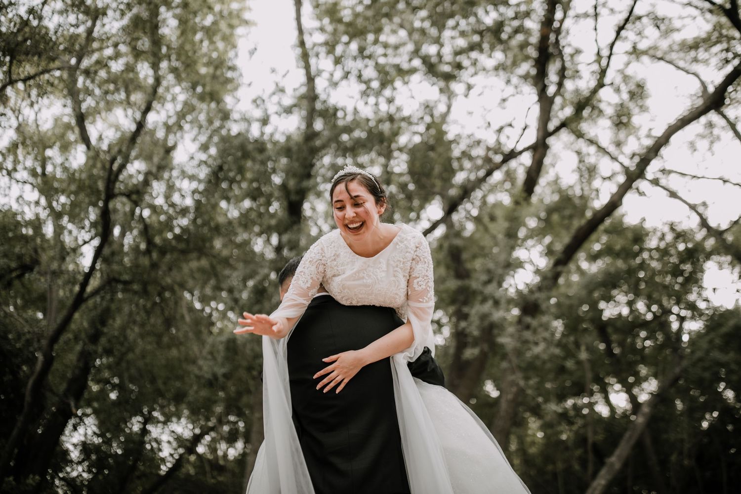 Joyful bride in lace wedding dress laughing outdoors surrounded by green trees.