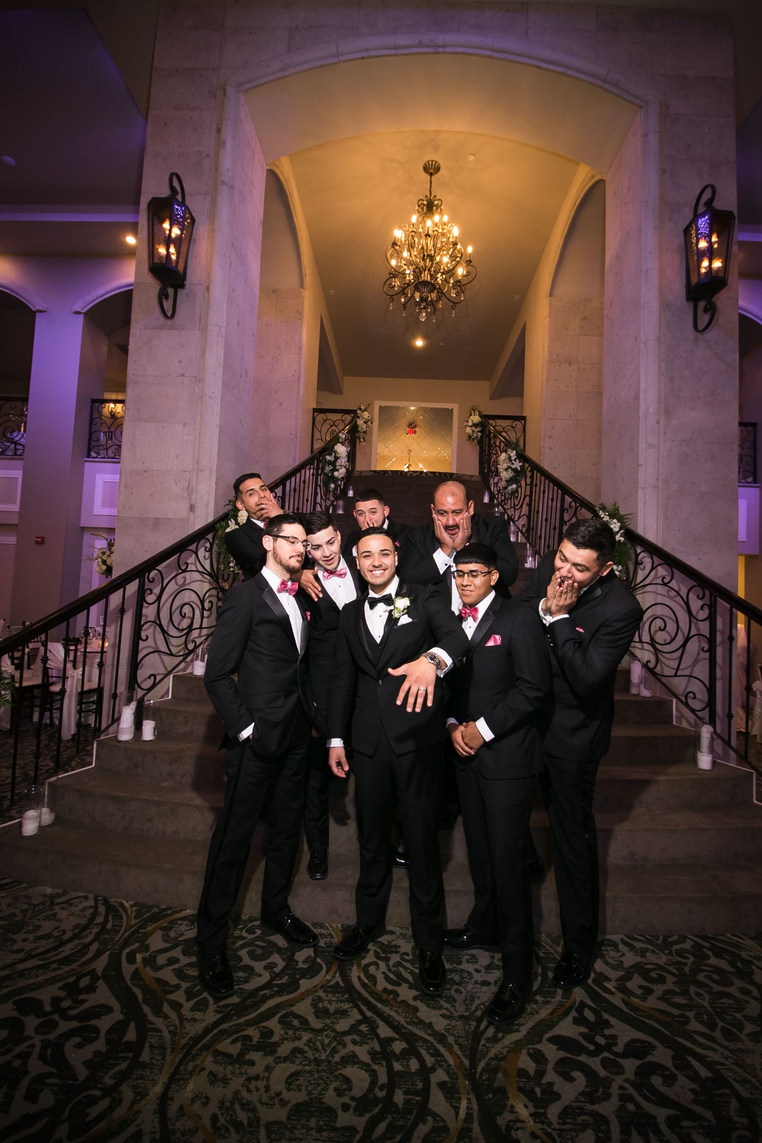 Groom and groomsmen in black tuxedos pose playfully on grand staircase with chandelier at wedding venue.