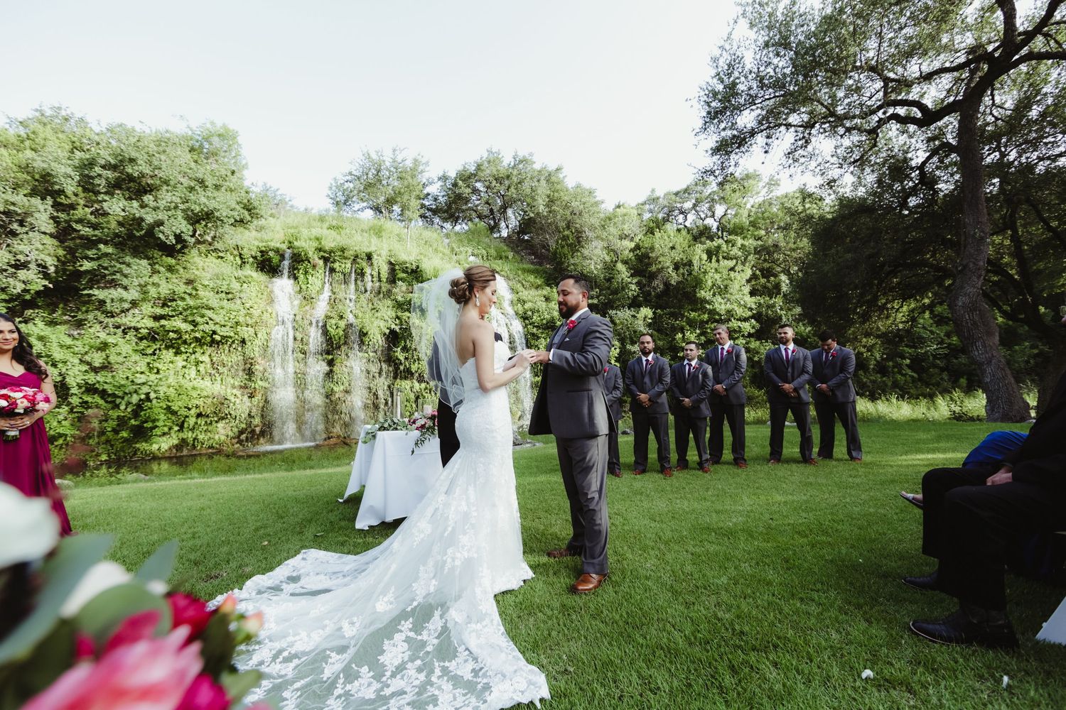 Bride and groom exchange vows at outdoor wedding ceremony with lush green backdrop.