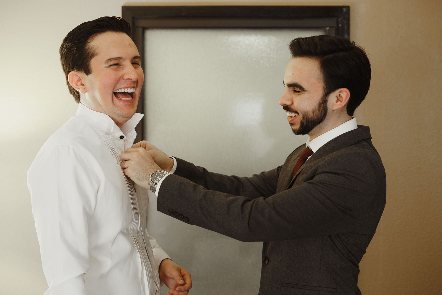 Groomsman helps laughing groom adjust his collar while getting ready for the wedding.