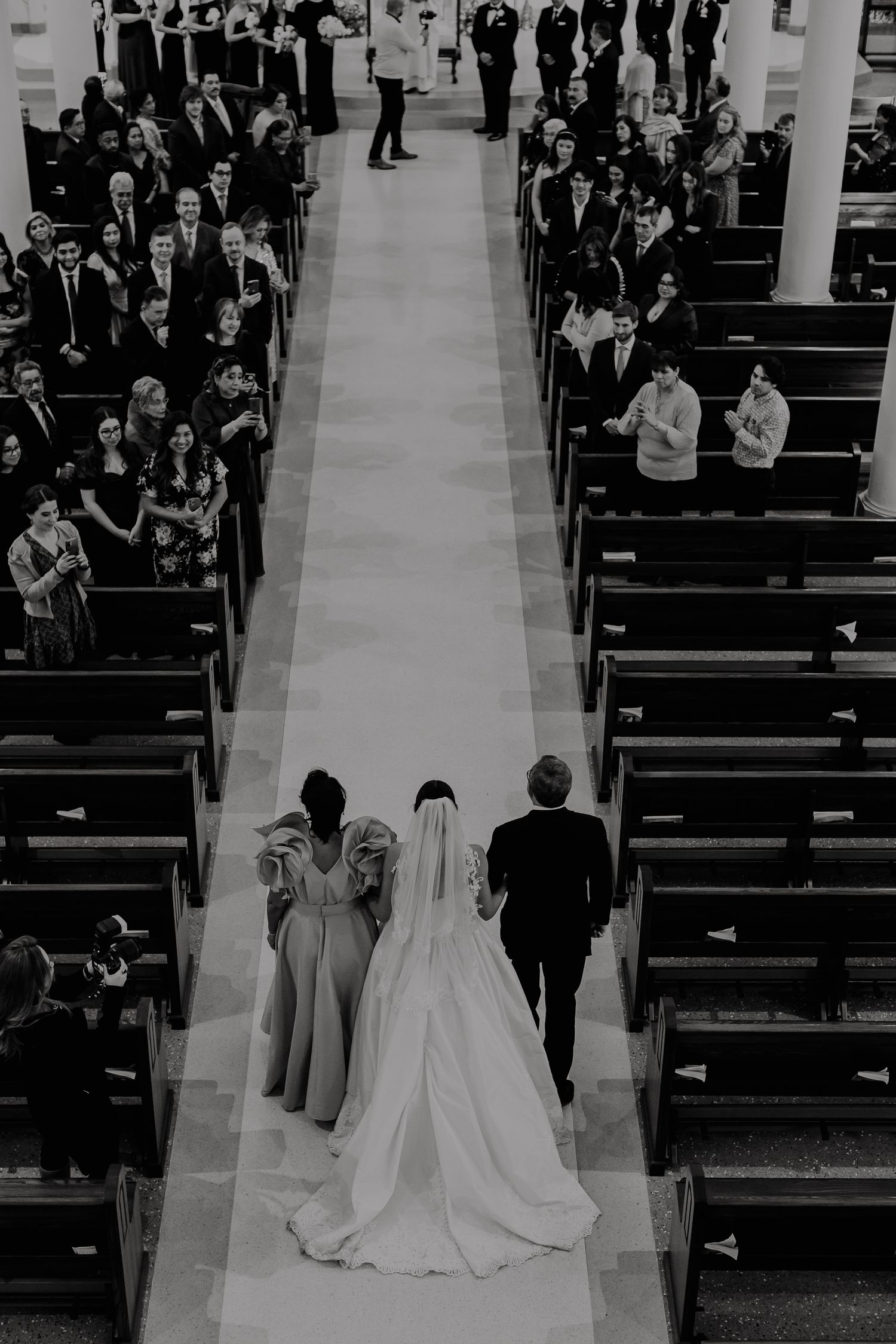 Black and white aerial view of a bride walking down the church aisle with wedding party, guests seated in pews.