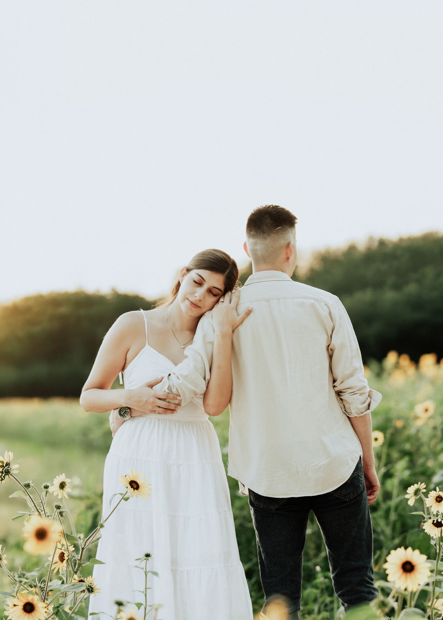 Couple embracing in sunflower field at golden hour, woman in white dress leaning on mans shoulder.
