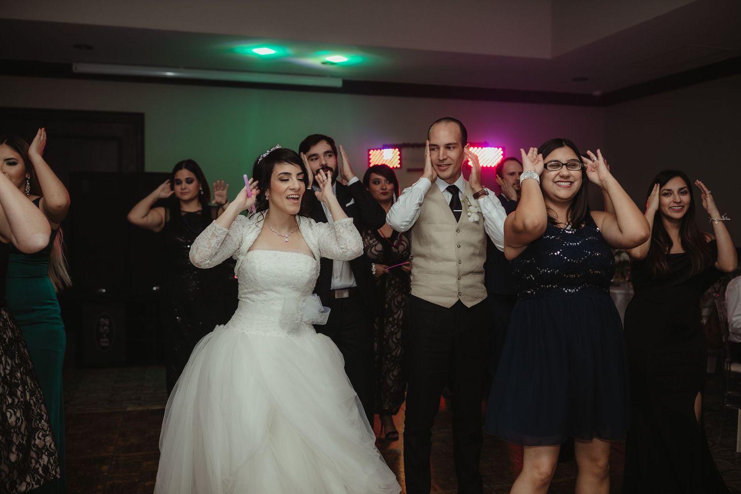 Bride and groom dancing joyfully with wedding guests on a colorfully lit dance floor.