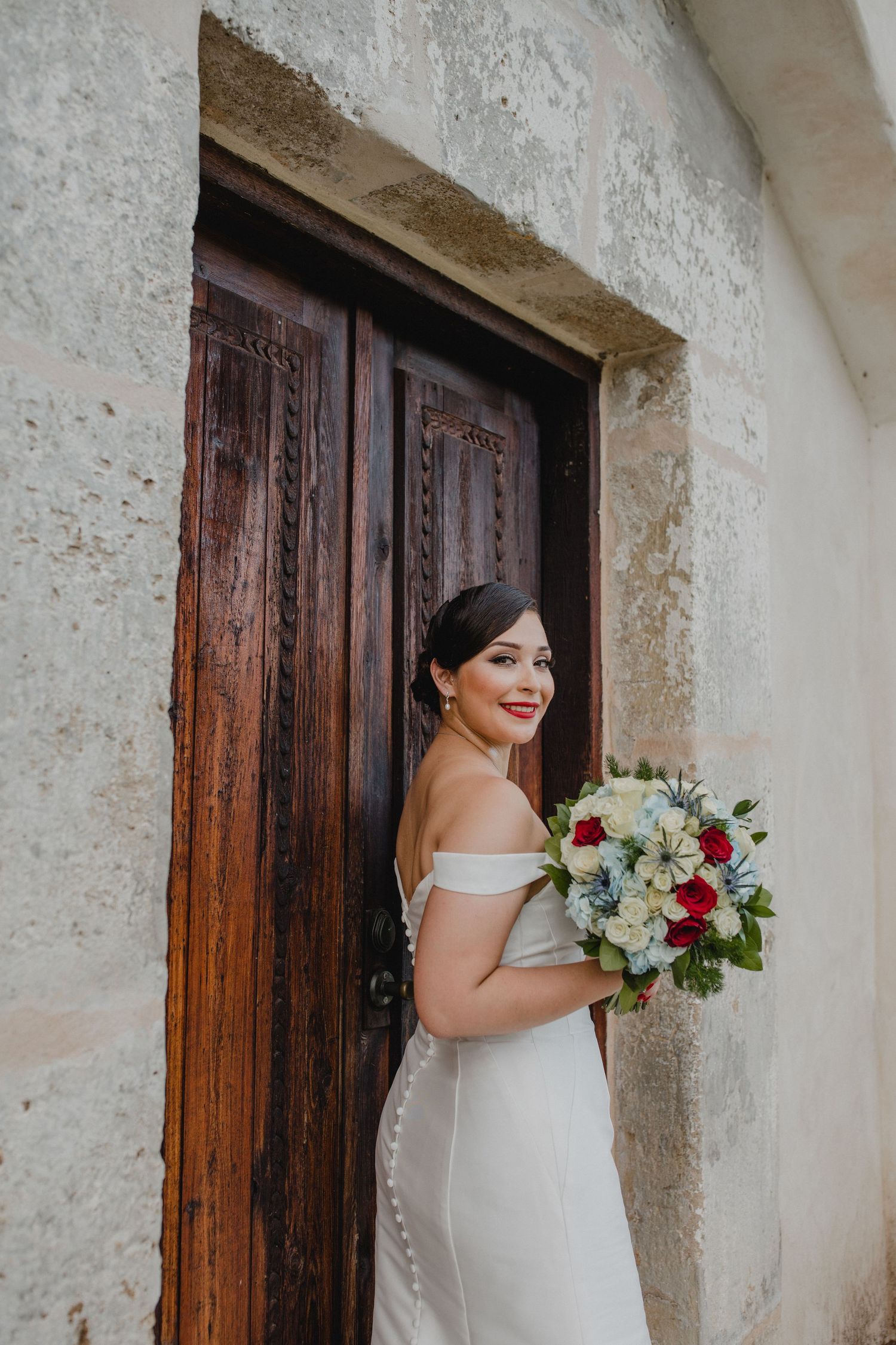 Smiling bride in an off-shoulder white gown holds a red and white bouquet beside a rustic wooden door.