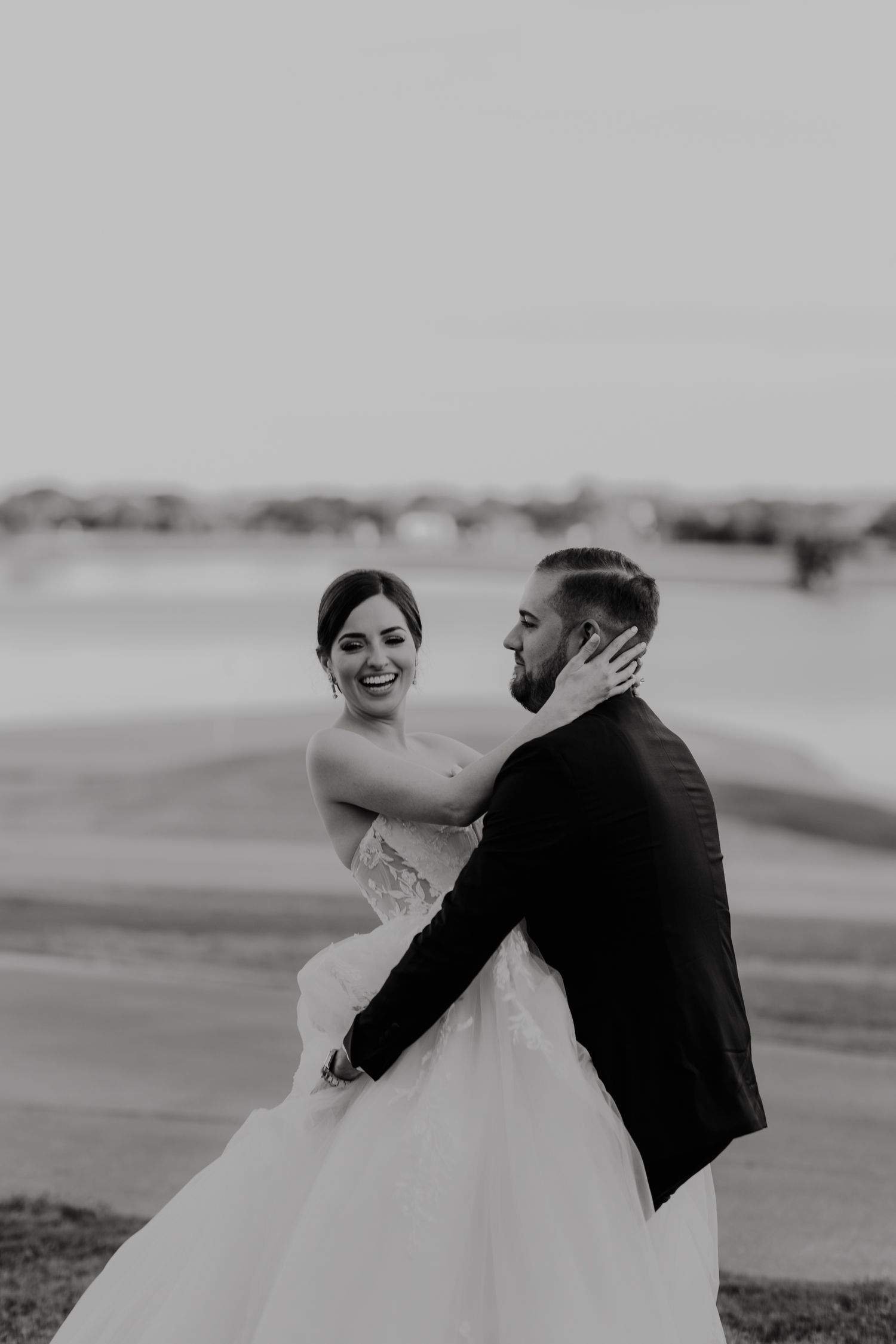 Black and white photo of a smiling bride and groom embracing outdoors on their wedding day.