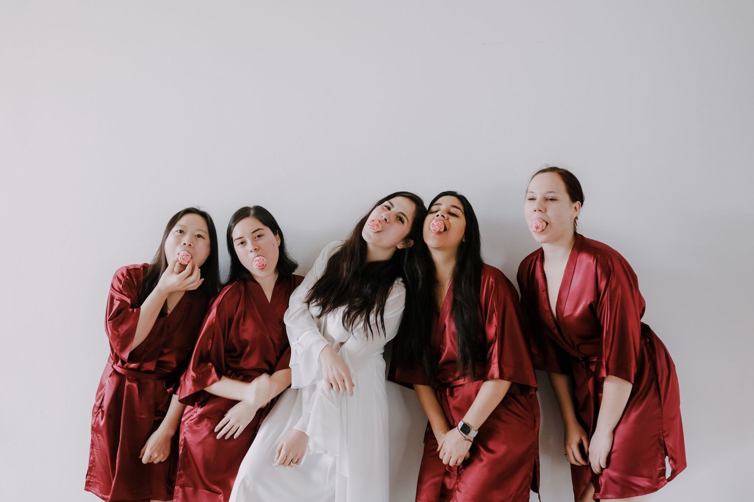 Bride and bridesmaids in burgundy silk robes make playful faces together against a white background.
