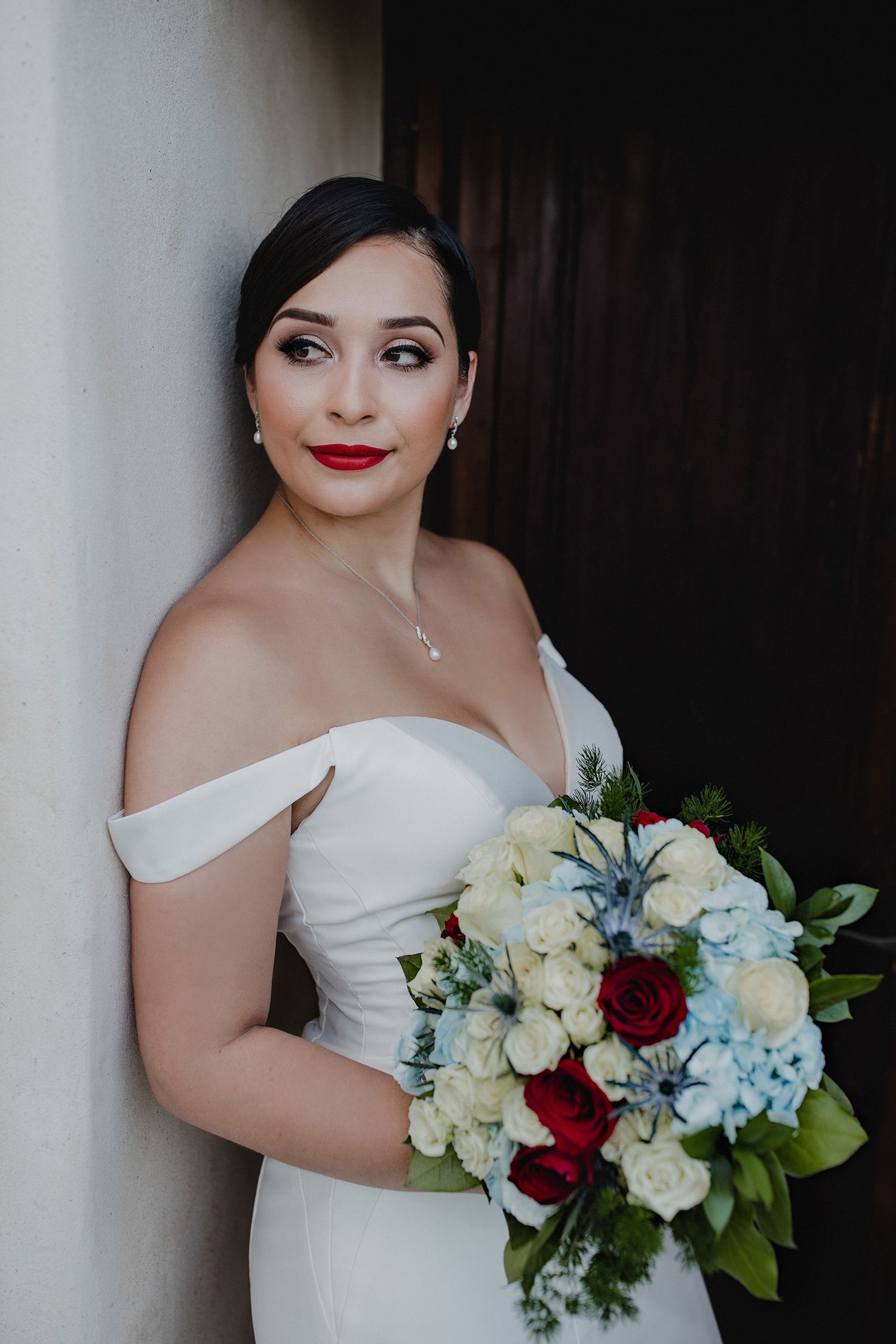 Beautiful bride in off-shoulder white gown holding a bouquet of red roses and blue hydrangeas.