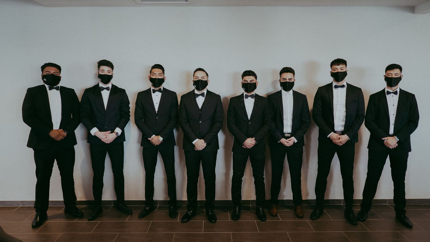 Eight men in black tuxedos and bow ties wearing face masks pose together for a formal group photo.