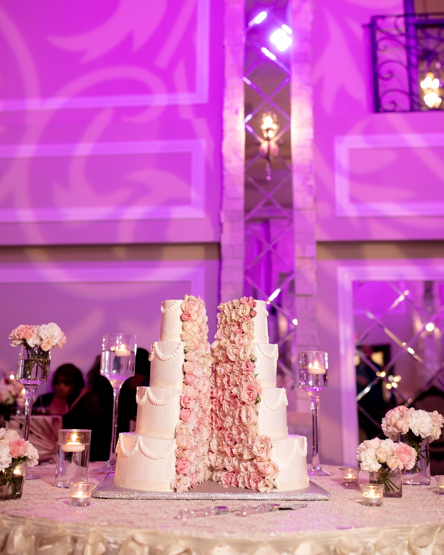 Elegant five-tier white wedding cake adorned with cascading pink roses under purple reception lighting.