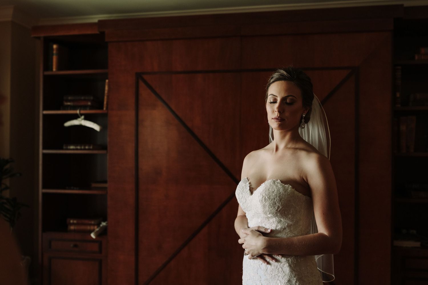 A bride in a strapless lace wedding gown stands in soft natural light near a wooden cabinet.