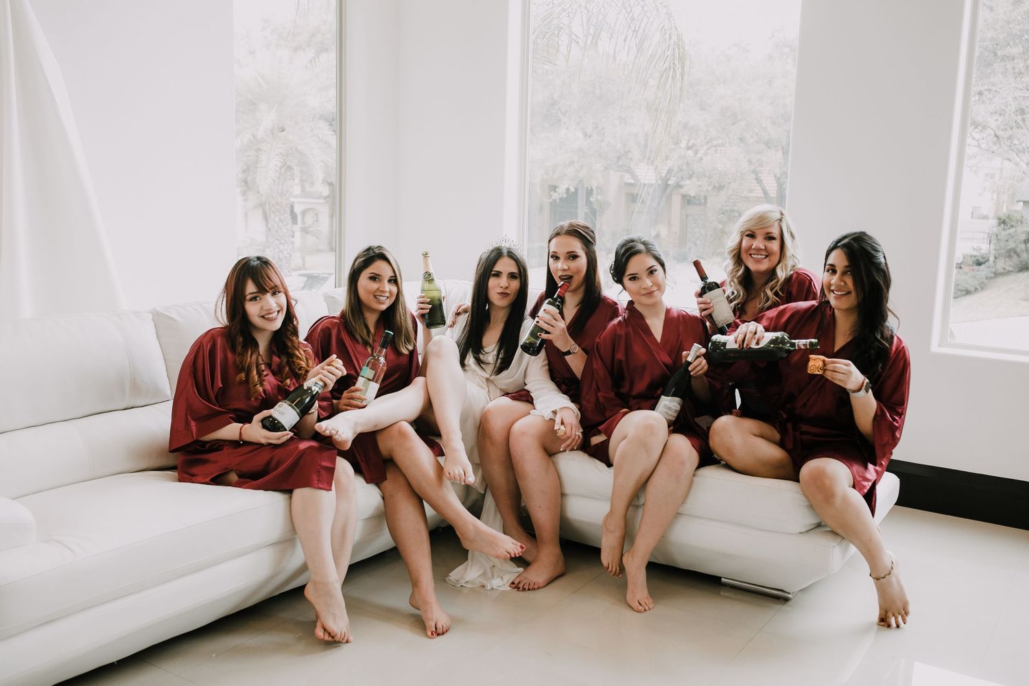 Bridesmaids in matching burgundy robes celebrate together on white sofa during wedding morning preparations.