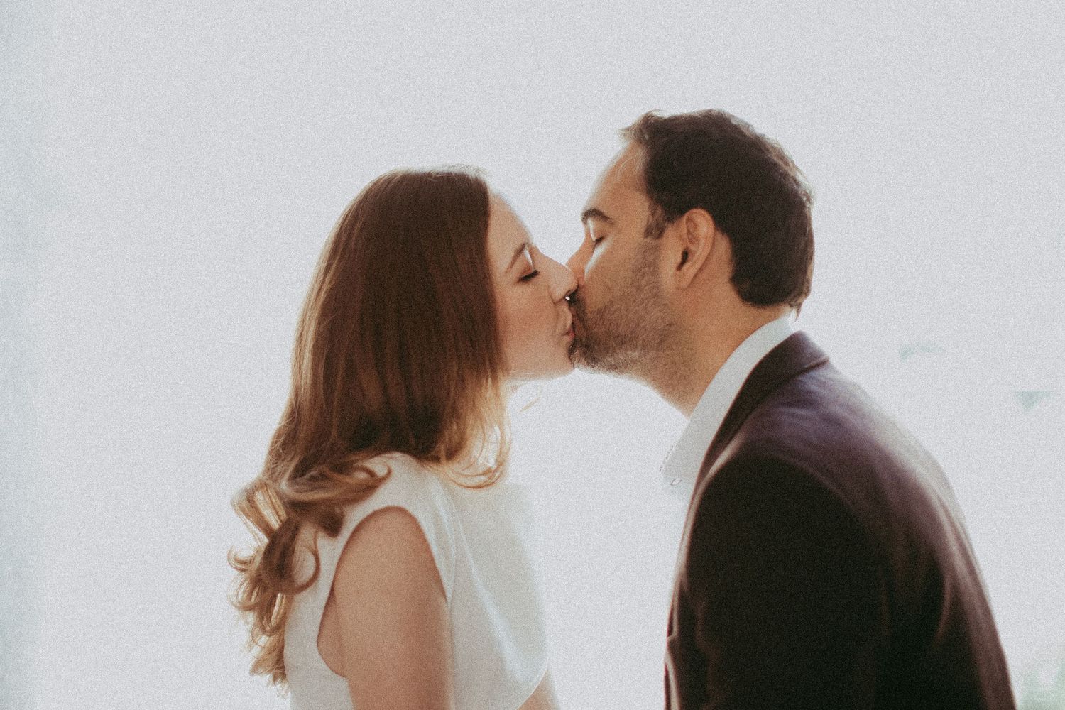 A couple sharing a romantic kiss, the woman in white and man in a dark suit, against a soft white background.