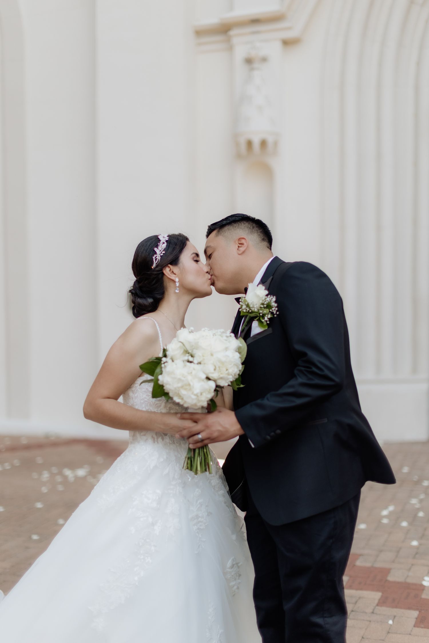 Wedding couple sharing a kiss while holding a white bouquet against a light architectural backdrop.