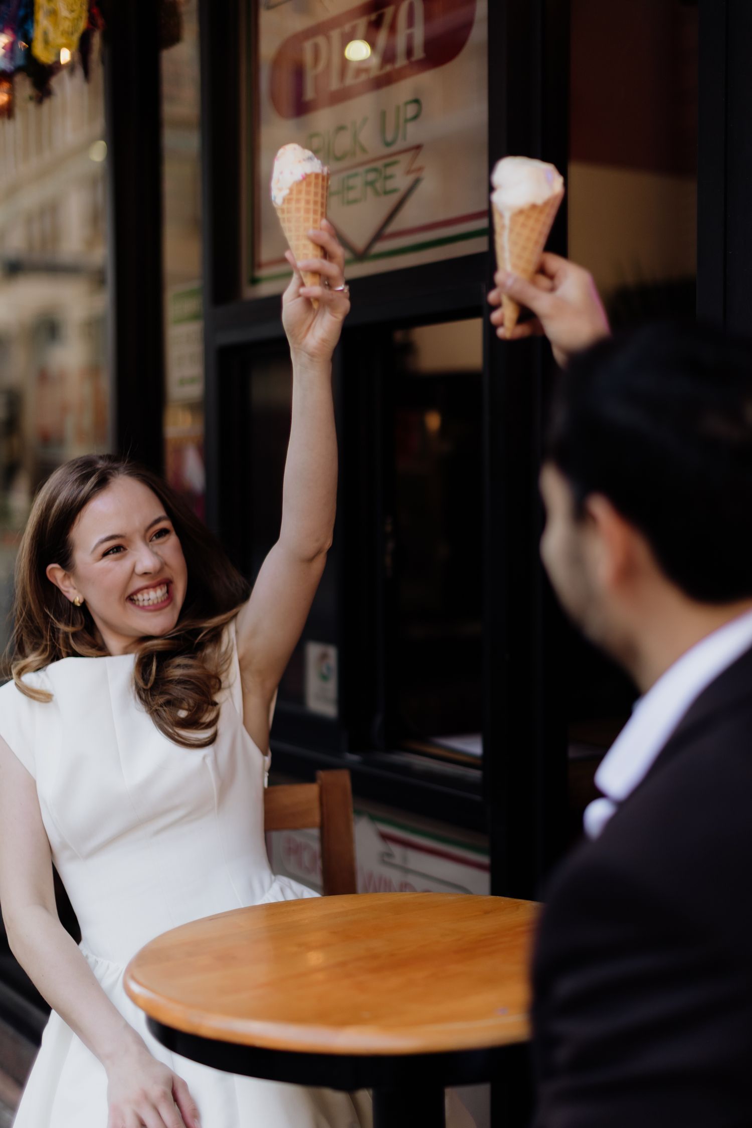 Joyful bride in white dress raising ice cream cone with groom at outdoor cafe table, celebrating together.