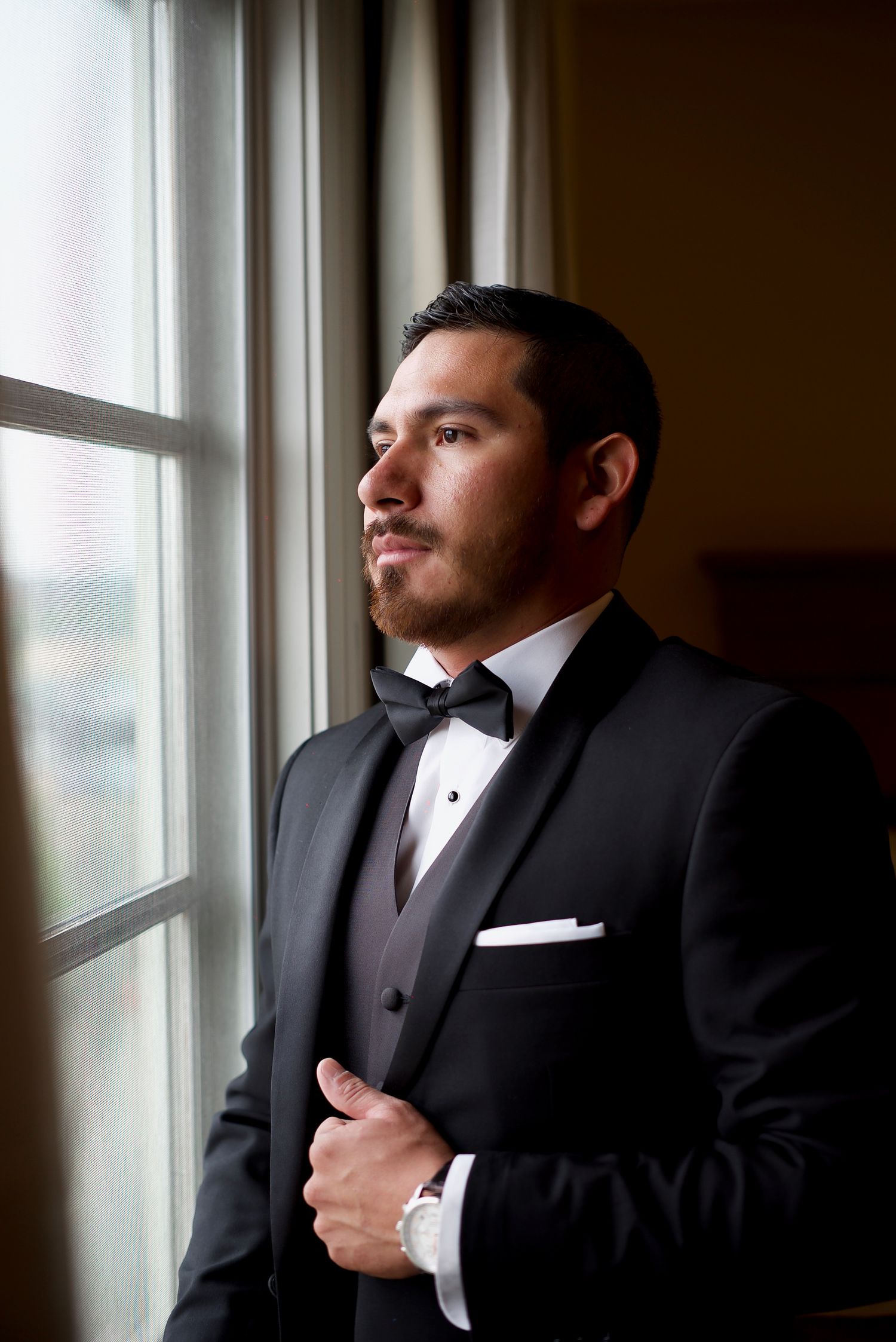 Elegant groom in black tuxedo gazing thoughtfully out window before wedding ceremony.