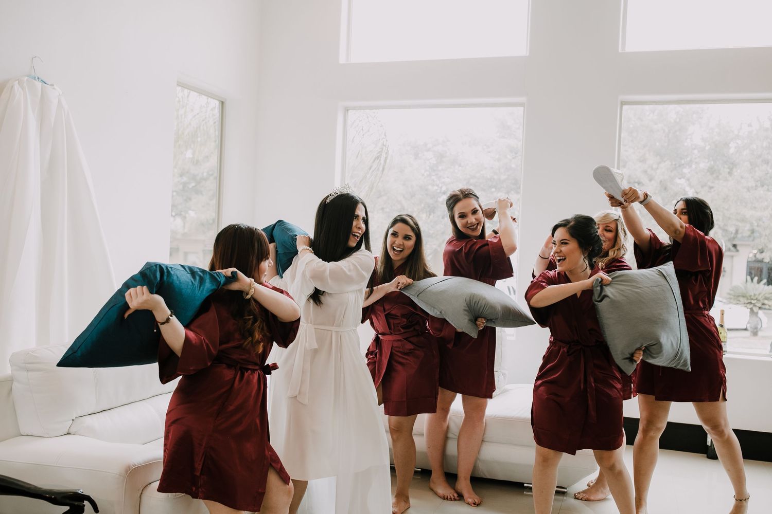 Bride and bridesmaids in burgundy robes enjoy playful pillow fight before wedding.