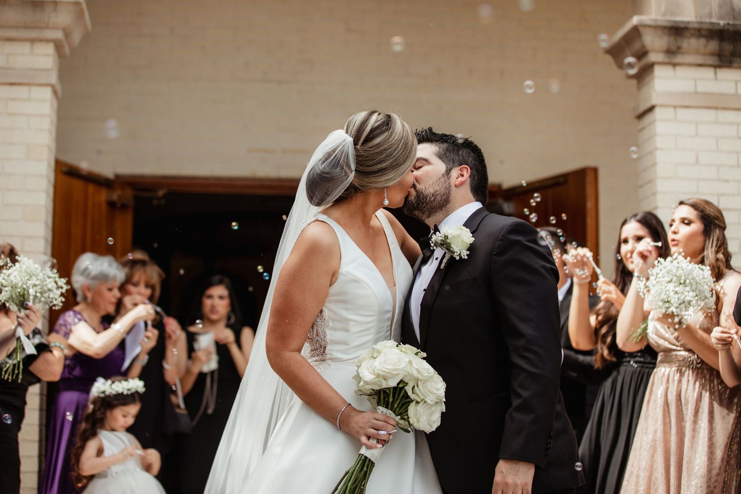 Newlyweds share romantic kiss surrounded by bubbles and cheering guests outside church after ceremony.