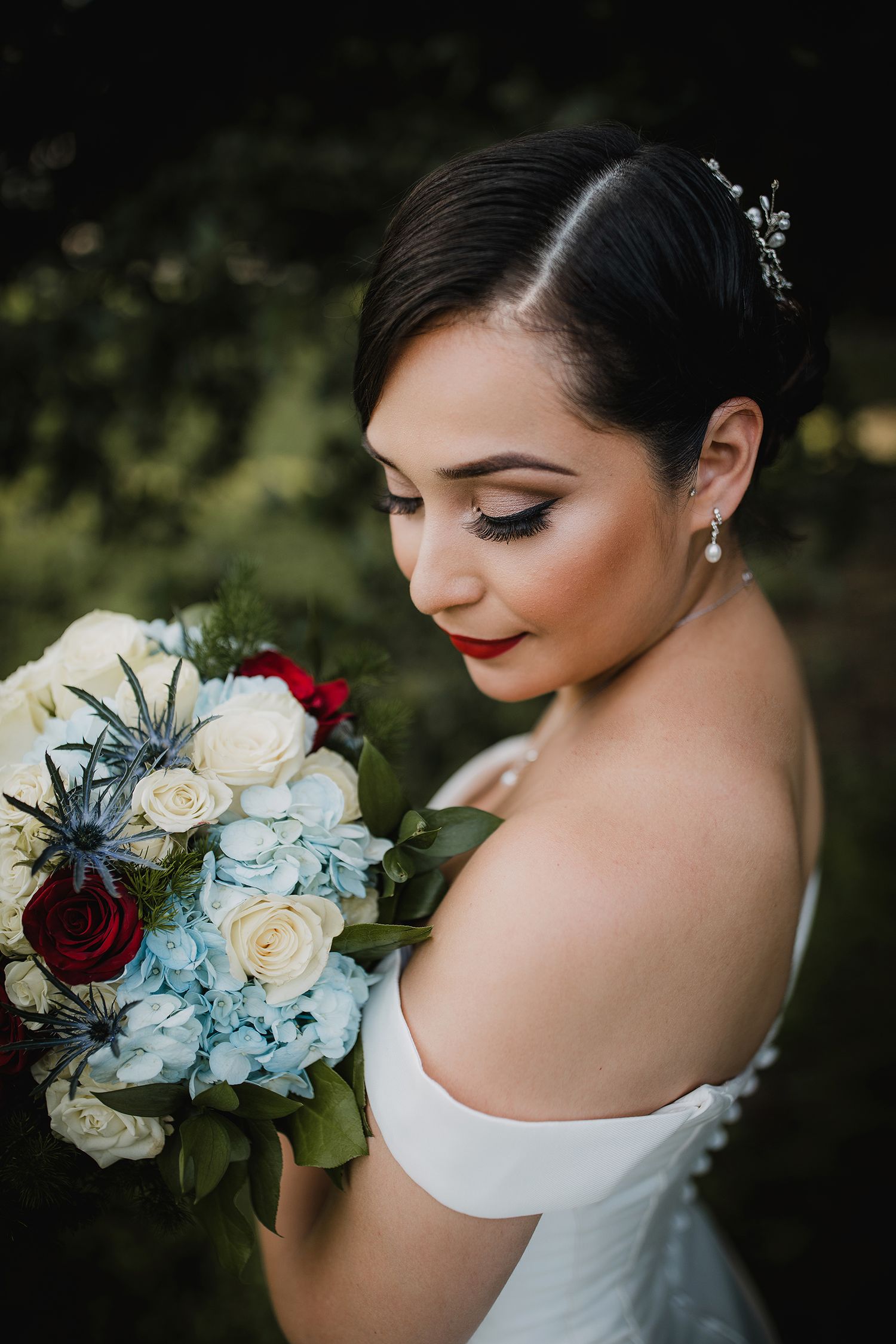 Elegant bride with updo hairstyle gazes down at bouquet of white roses, red flowers, and blue hydrangeas.
