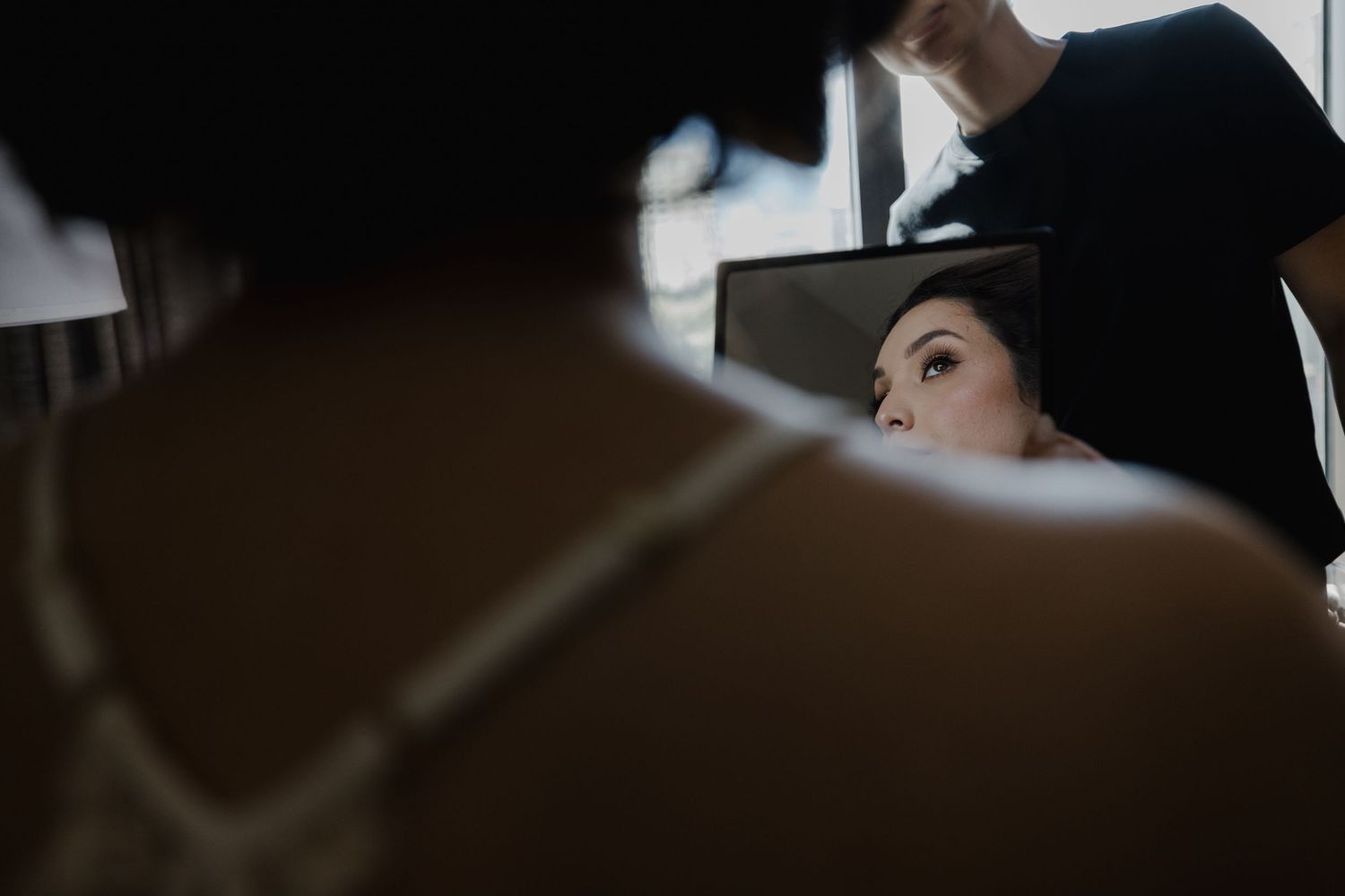 Bride getting hair styled, reflected in mirror during wedding preparation, captured in moody dark tones.