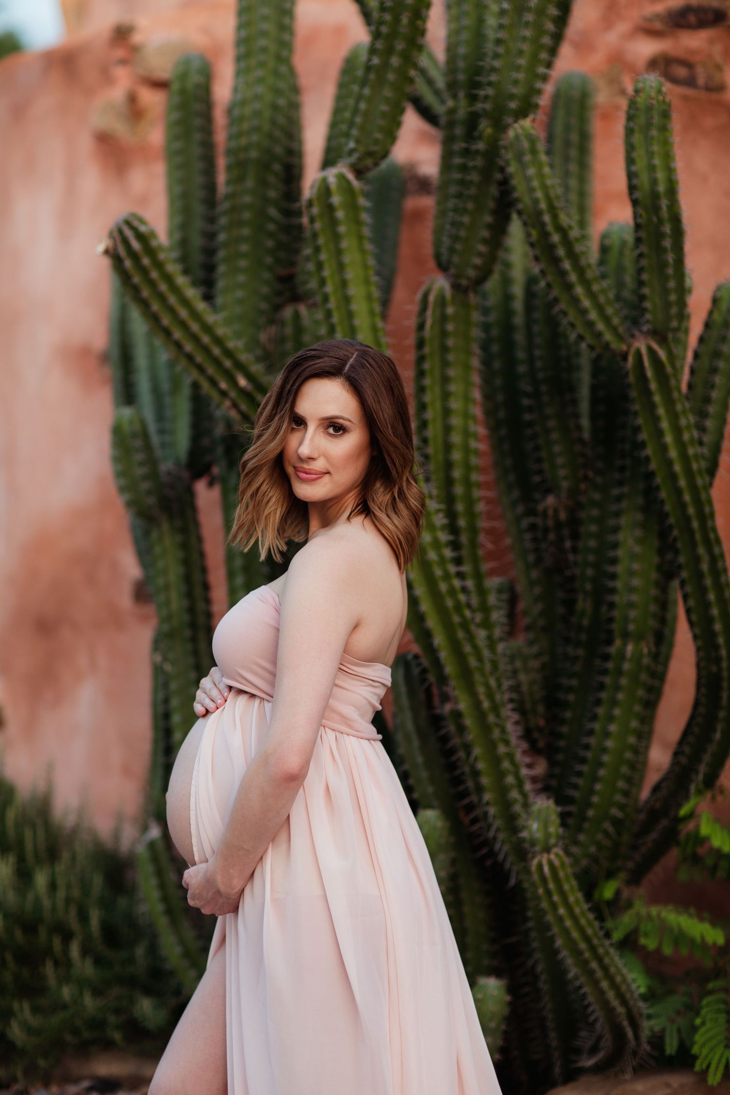 Artistic maternity portrait with dramatic cactus garden and pink adobe wall backdrop.