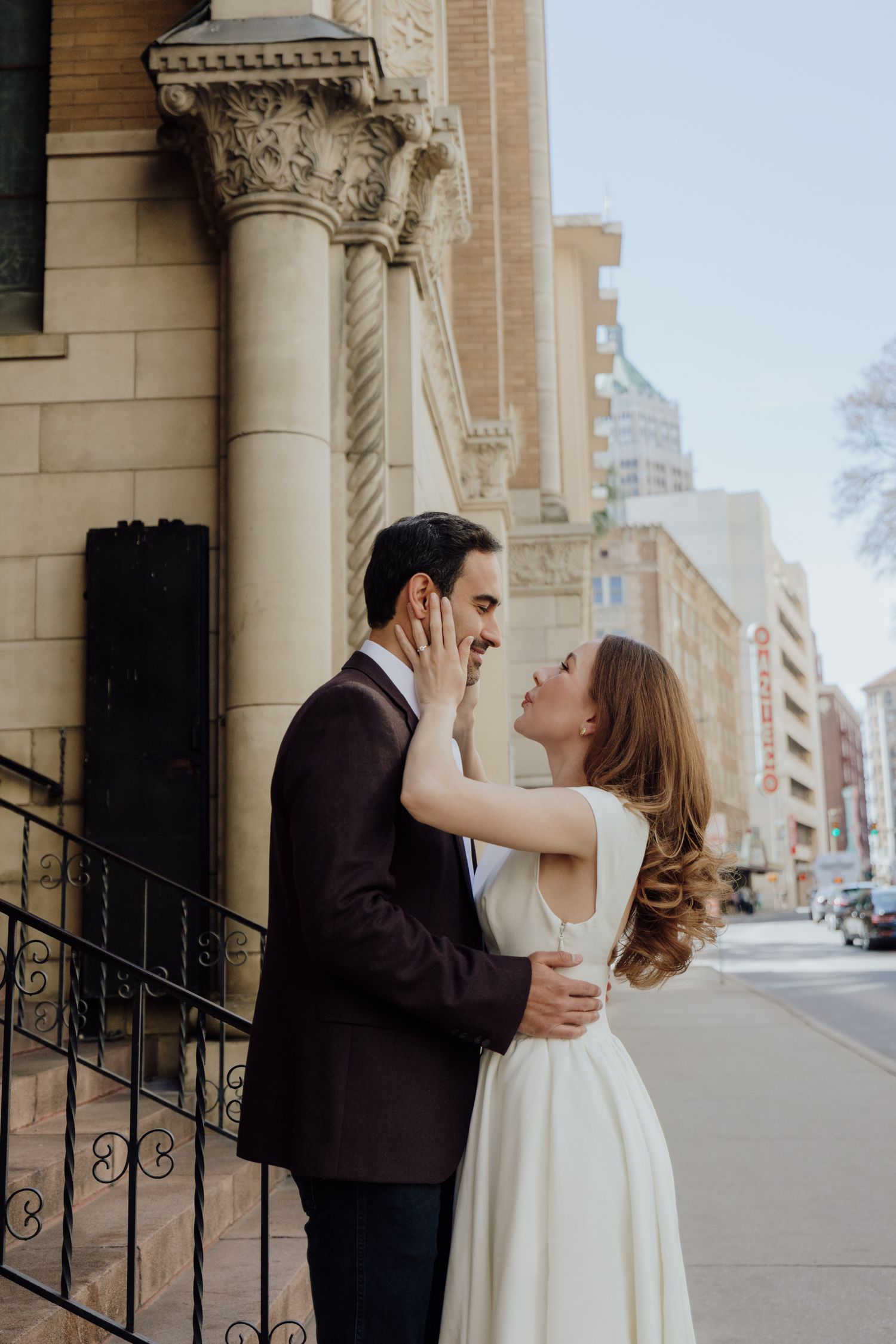 A couple in wedding attire embraces and smiles on a city sidewalk beside a historic stone building.