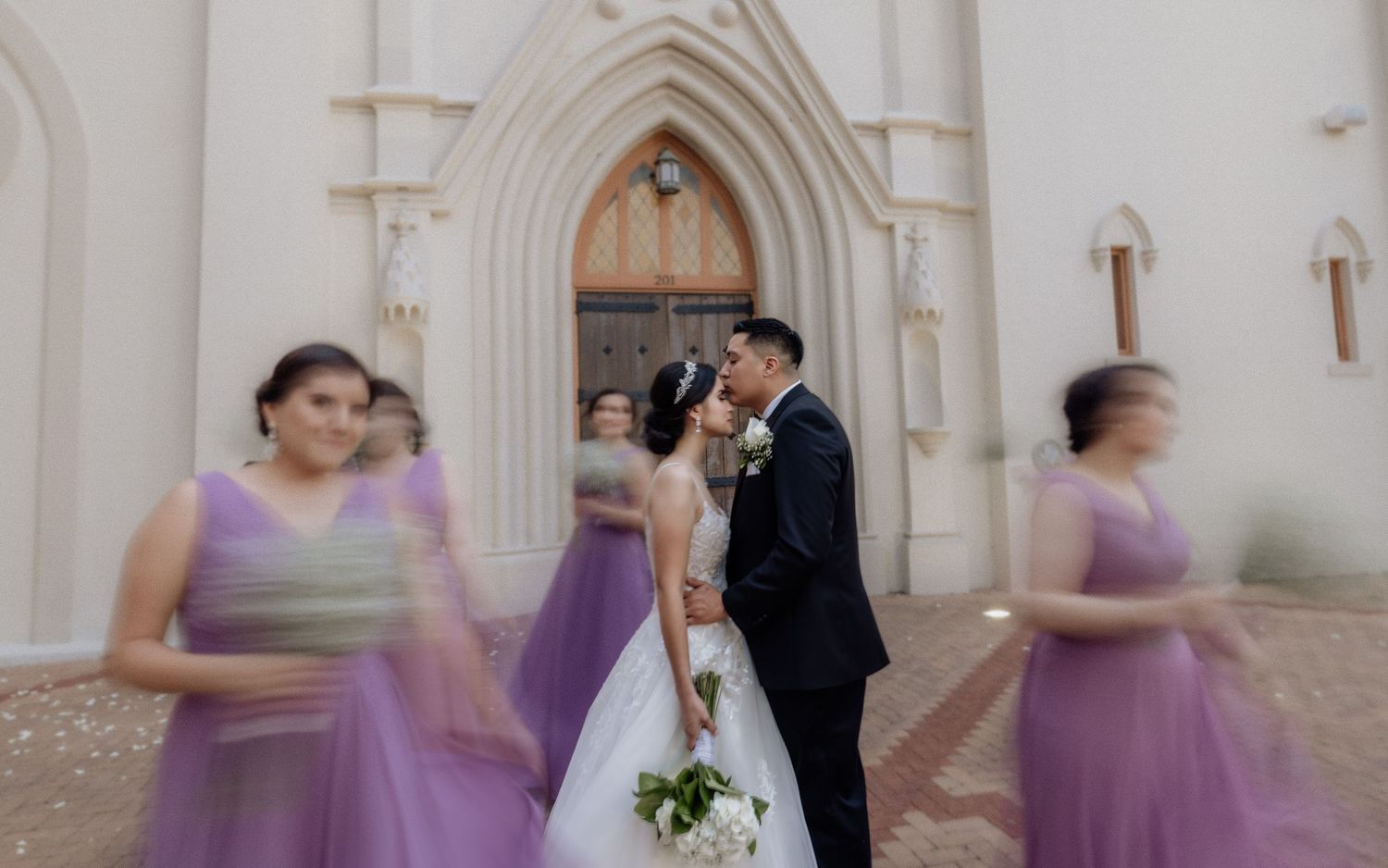 Bride and groom kissing outside a white church, surrounded by bridesmaids in purple dresses on their wedding day.