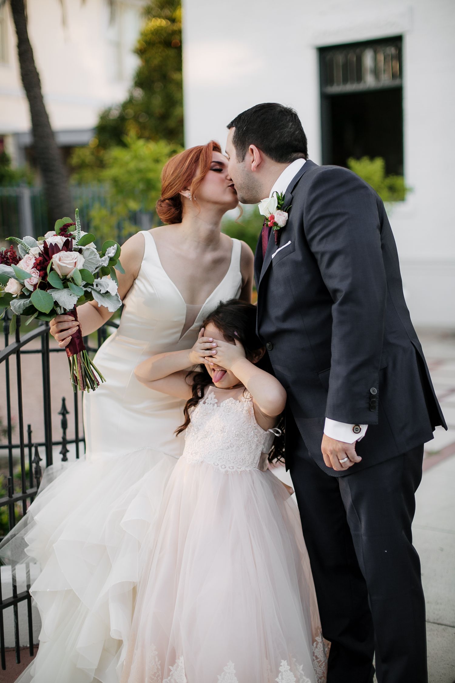 Bride and groom kissing while flower girl covers her eyes on wedding day.