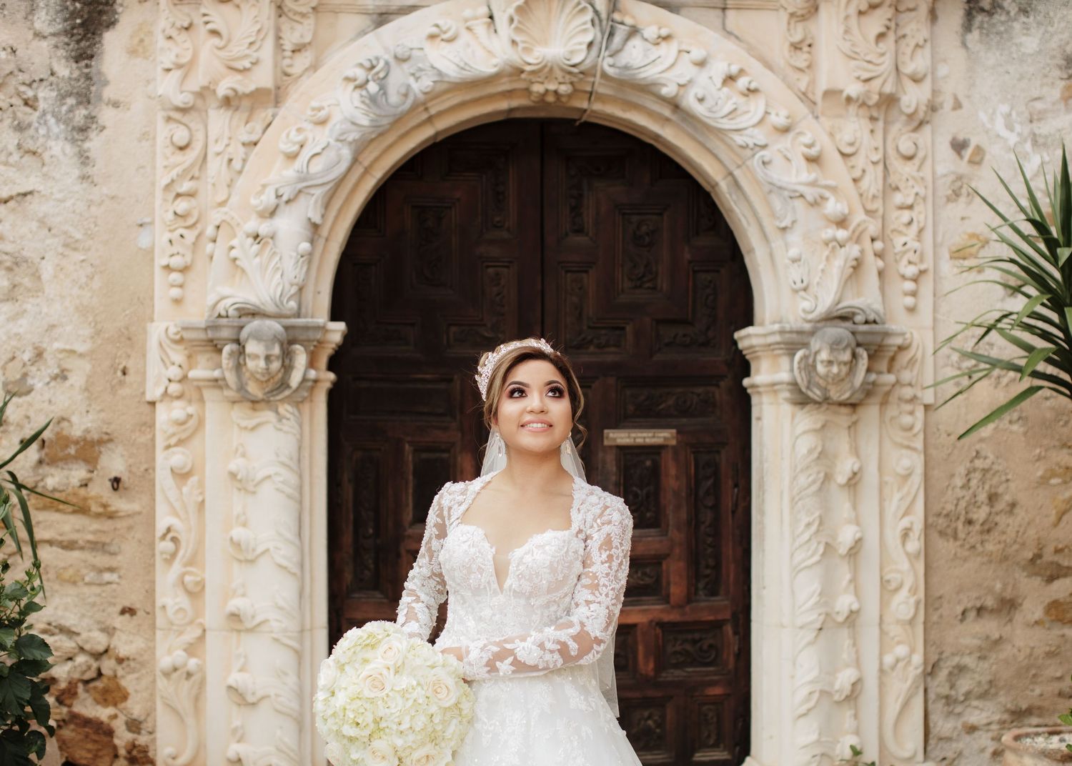 Stunning bride in lace gown and veil poses under ornate stone archway at historic venue.