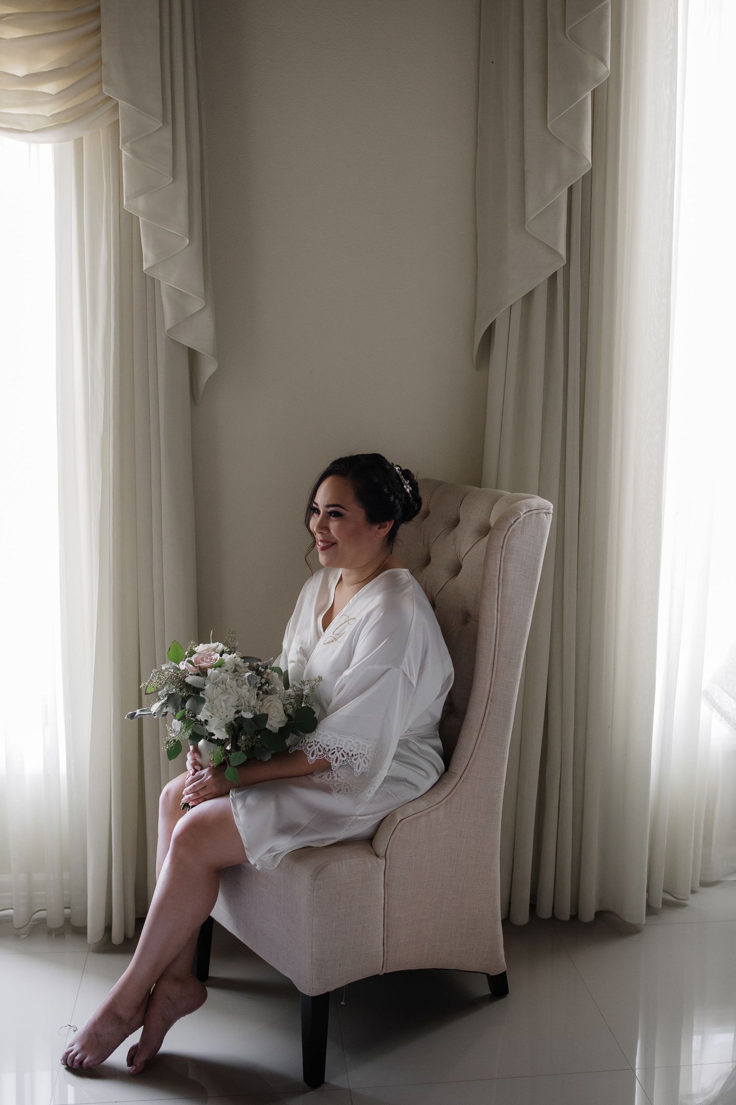 Bride in white robe sits in elegant chair holding bouquet near bright curtained window.