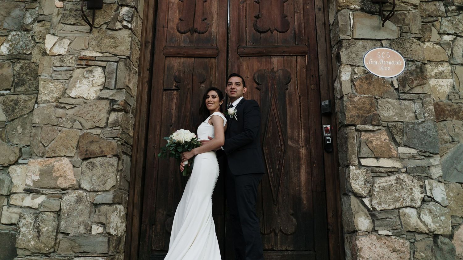 Bride and groom posing together in front of rustic stone church with large wooden doors on wedding day.