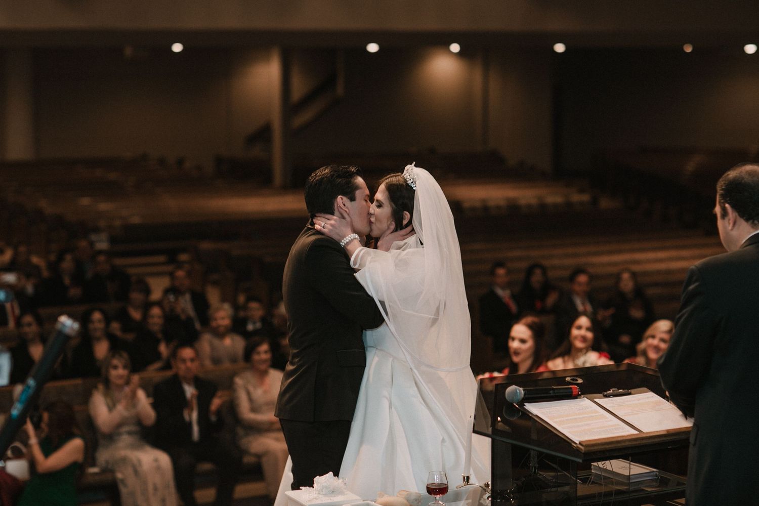 Bride and groom share their first kiss at the altar before a large audience in a grand wedding venue.