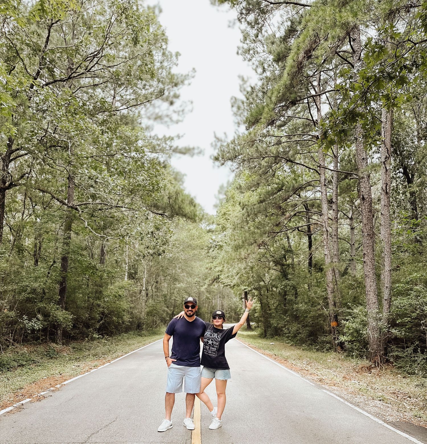 Two people stand together in the middle of a tree-lined road through a lush green forest.