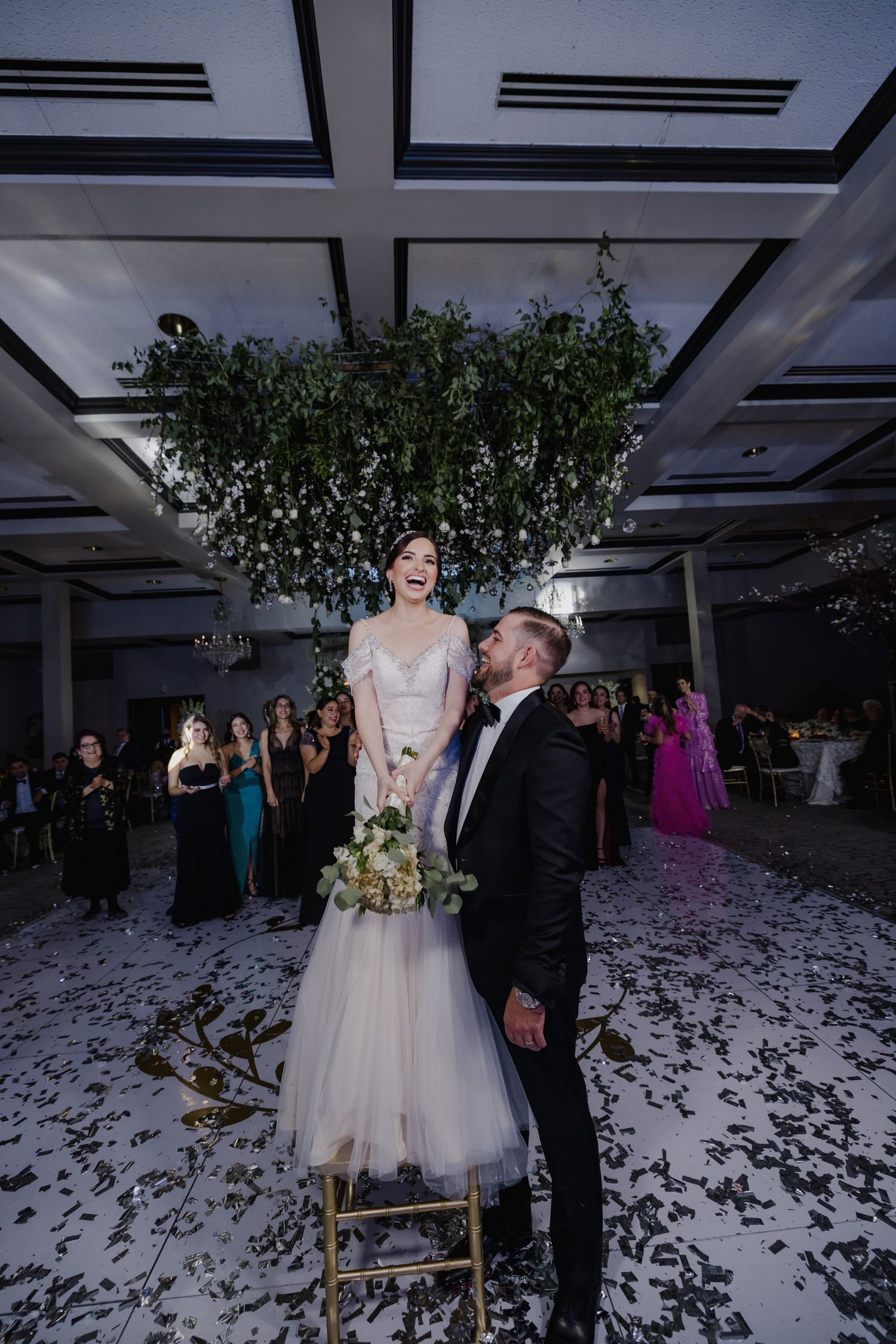 Bride laughing on chair while groom kisses her cheek on confetti-covered wedding reception dance floor.