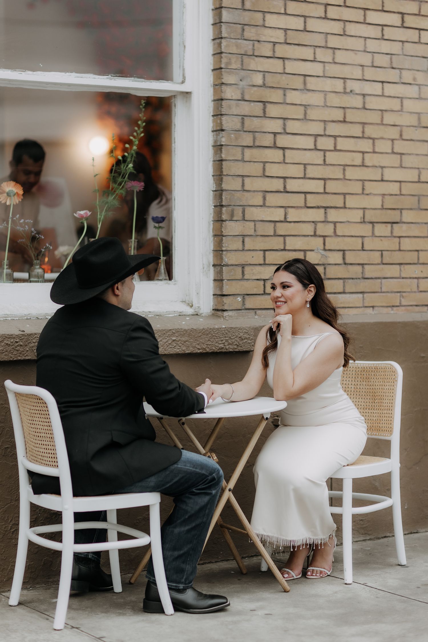 Couple in wedding attire sitting at a white bistro table outside a brick building, bride smiling in white dress.